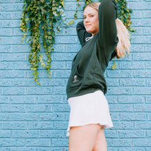 Load image into Gallery viewer, Model wearing green sweatshirt and white skirt in front of a blue wall 
