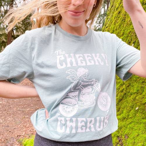 girl wearing light blue t shirt in the forest. The t shirt features a cherub riding a motorcycle and says 'The Cheeky Cherub'