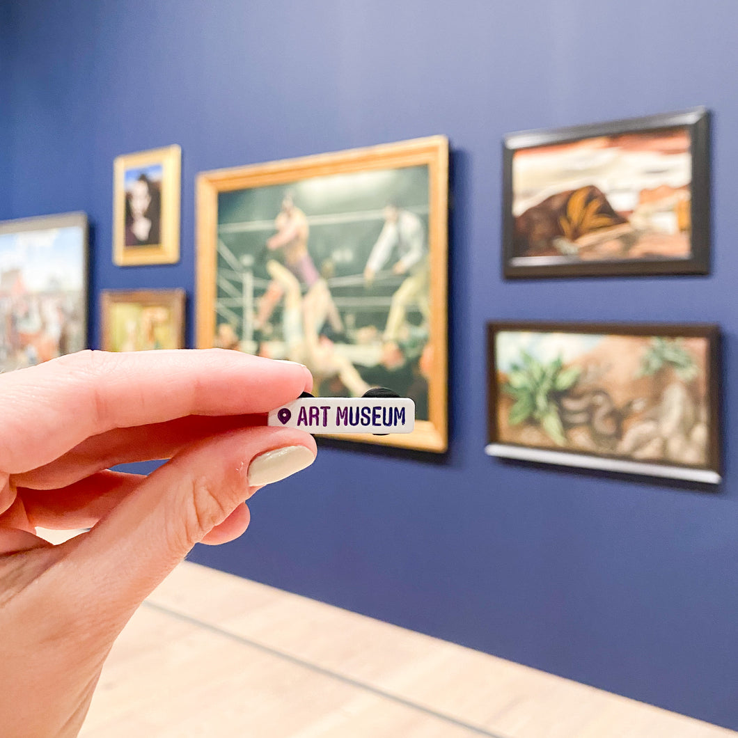 Hand holding a pin with the Instagram location symbol and the world 'art museum' in front of a blue wall filled with paintings