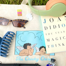 Load image into Gallery viewer, Tote bag with two cherubs, sunglasses, enamel pins, a book, and over the ear headphones
